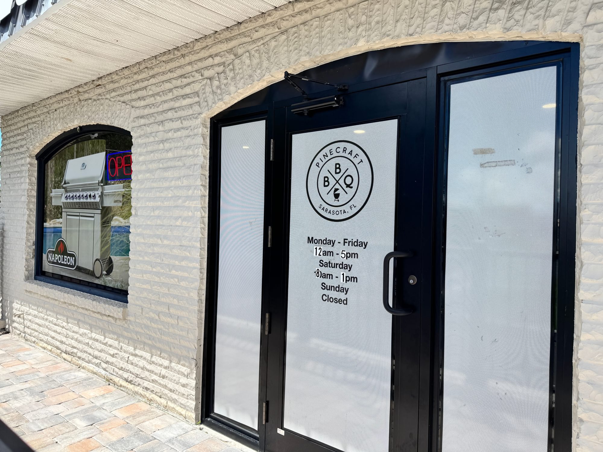 Glass entrance door with the round Pinecraft Barbecue logo and posted hours.