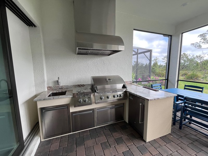 Covered outdoor kitchen with a grill and hood beside a table and chairs.